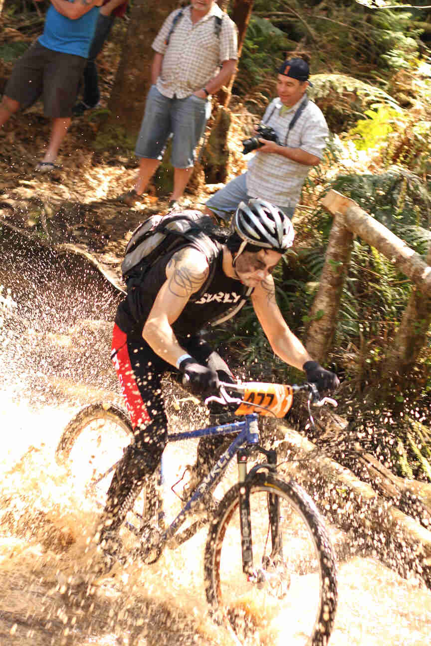 Front, right side view of a cyclist splashing through water, as they ride their blue Surly bike in a forest